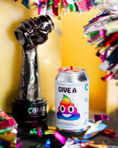 Photo of "Give a Crap" beer can with the craft beer marketing award behind. Can is white with text "Give A" and a smiling rainbow poop emoji below. Craft Beer Marketing award is a platinum trophy of a hand directed upwards and crushing a beer can in the fist. The background is golden yellow with shiny rainbow confetti around the award and beer can. 