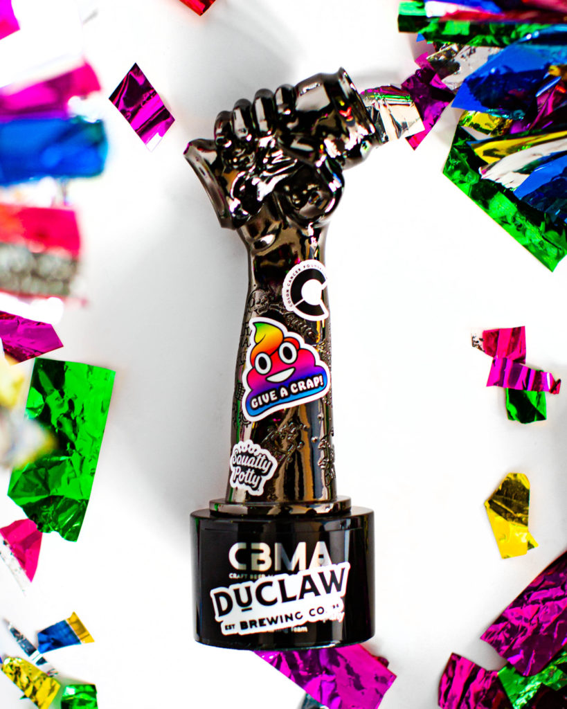 Image of Crushie trophy for beer marketing award: a fist crushing a beer can, laid flat on a white background with rainbow confetti. Trophy has various "Give A Crap" themed stickers stuck to it.