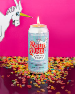 Image of the DIY Sour Me Unicorn Farts beer can candle. Can is against a pink background with fruity cereal spread around. A white unicorn is peeking out from the left corner.